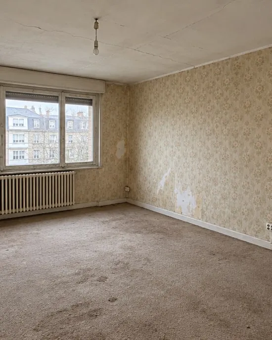 Séjour avant travaux : papier peint déchiré, moquette usée, plafond fissuré et prises en saillie