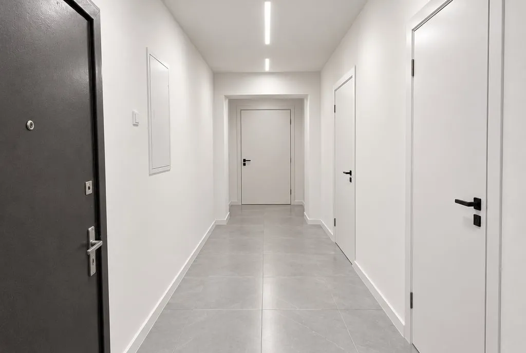 Couloir d'entrée après travaux : murs blancs lisses, grand carrelage gris, tableau encastré et éclairage LED