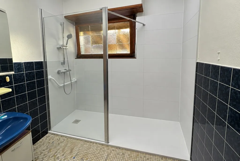 Salle de bain après travaux : douche à paroi vitrée sous le vitrail, faïence blanche, barre d'appui