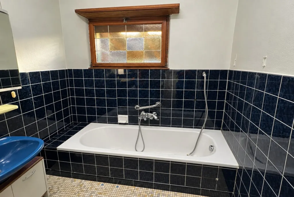 La même salle de bain avant travaux : baignoire encastrée sous le vitrail, carrelage bleu marine