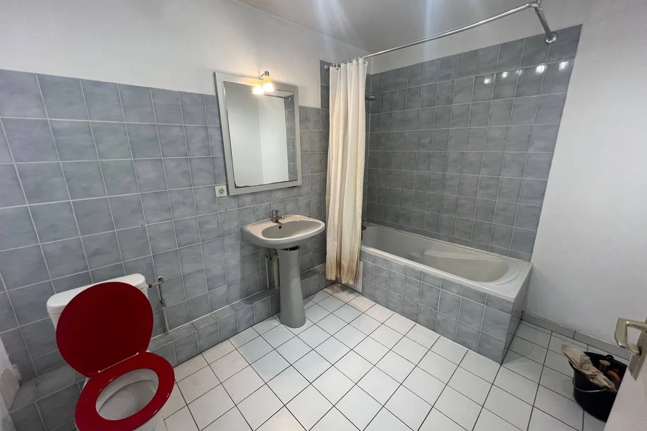 La même salle de bain avant travaux : carrelage gris, lavabo sur colonne, baignoire à rideau et WC à abattant rouge